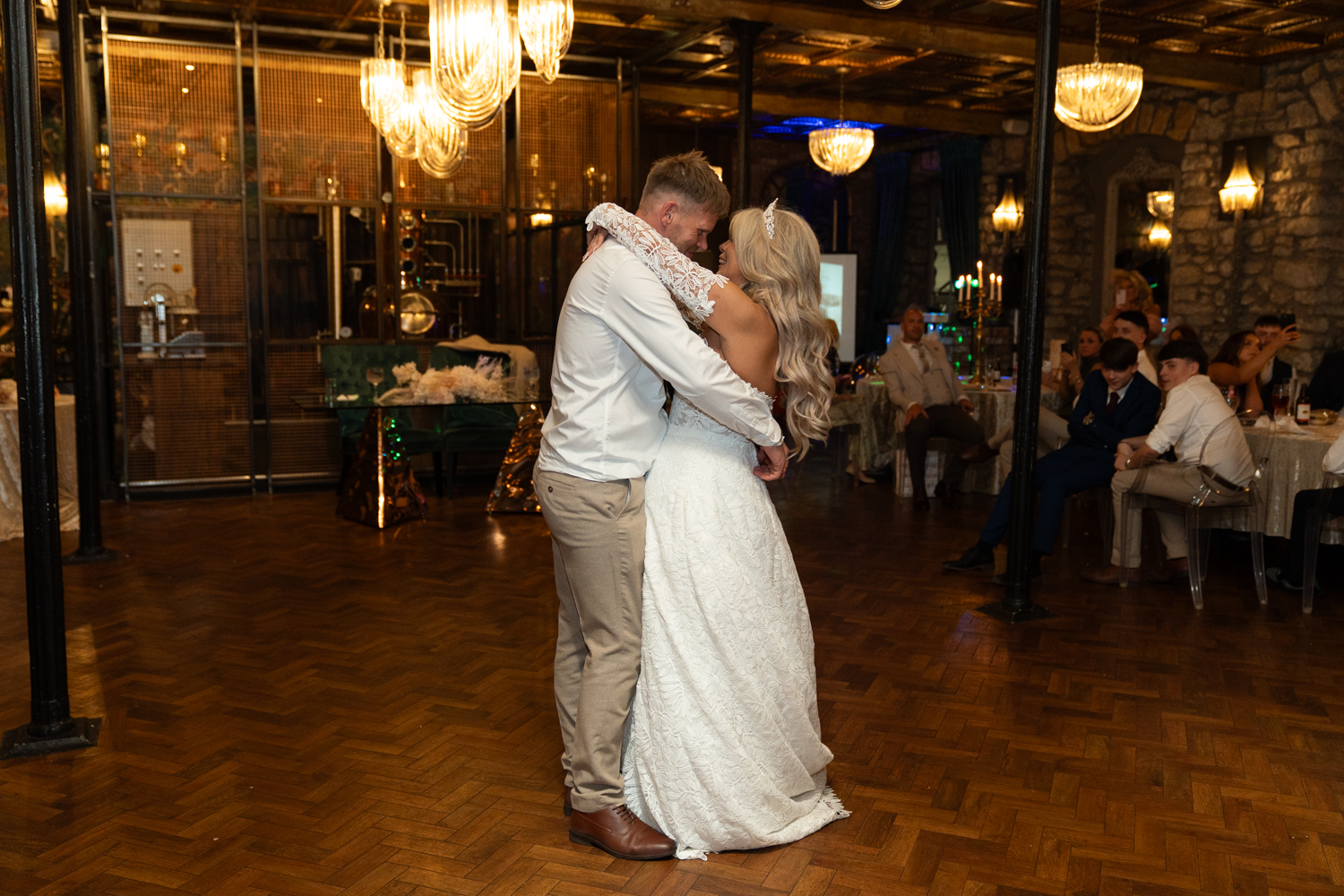 The couple dancing together, seen from across the room — guests watching from their seats, the venue glowing under warm light